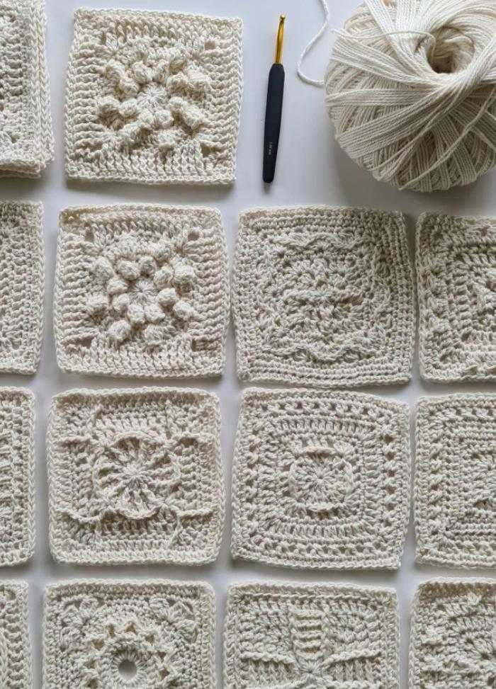 A collection of intricately crocheted white squares, each with unique patterns, arranged neatly. A crochet hook and yarn ball are visible, suggesting crafting in progress.