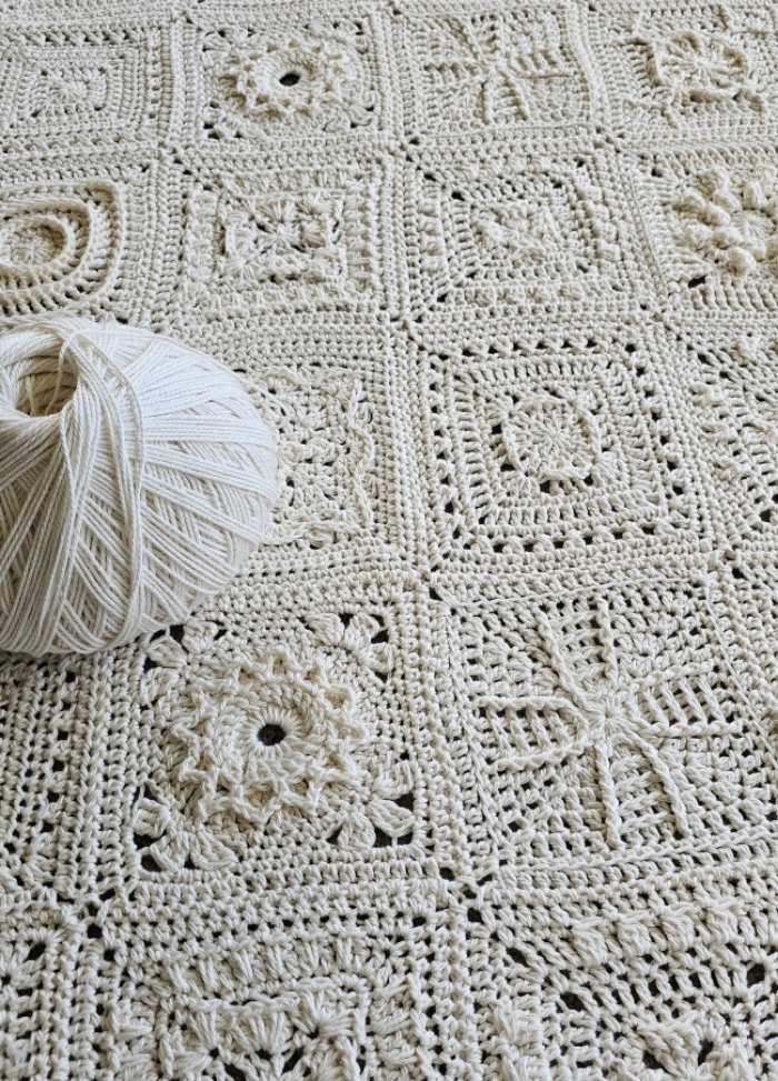 A detailed crochet blanket made from cream-colored yarn squares, each with unique patterns. A yarn ball sits on top, showcasing intricate craftsmanship.