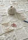 A ball of cream yarn sits on a table amidst a grid of crocheted squares. Red scissors and a blue crochet hook are placed nearby, conveying creativity.