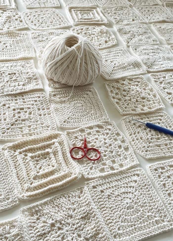 A ball of cream yarn sits on a table amidst a grid of crocheted squares. Red scissors and a blue crochet hook are placed nearby, conveying creativity.