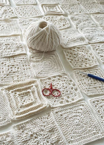 A ball of cream yarn sits on a table amidst a grid of crocheted squares. Red scissors and a blue crochet hook are placed nearby, conveying creativity.