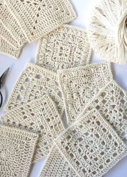 A collection of beige crochet squares and a ball of yarn on a white background. Scissors are nearby, suggesting a crafting session.