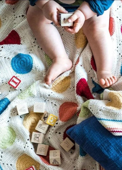 A baby sits on a colorful crocheted blanket with large polka dots, holding a block, surrounded by scattered alphabet blocks, evoking a playful and cozy atmosphere.