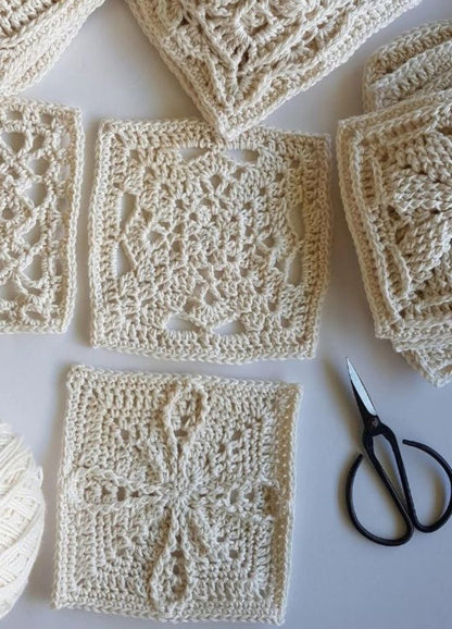 Cream crochet squares with intricate patterns are arranged on a white surface, surrounded by scissors and a ball of yarn, suggesting a calm, creative crafting scene.