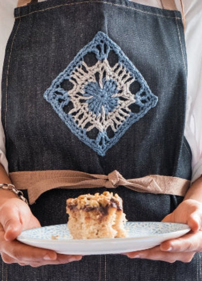 Person holding a blue-patterned plate with a piece of cake, wearing a denim apron featuring a crocheted blue and white diamond pattern. Cozy, homemade vibe.