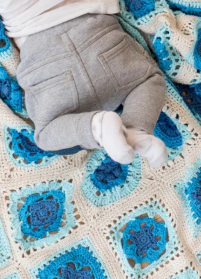 A baby in gray pants and white socks lies on a crocheted blanket with blue and white squares. The scene feels cozy and peaceful.
