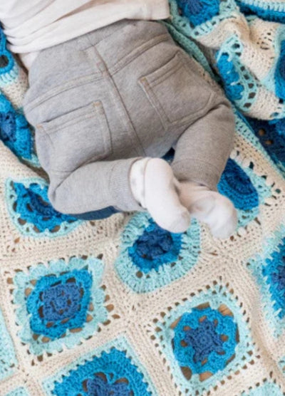 A baby in gray pants and white socks lies on a crocheted blanket with blue and white squares. The scene feels cozy and peaceful.