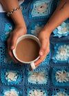 Hands holding a mug of coffee rest on a blue crocheted blanket. The blanket features intricate square patterns, creating a cozy, comforting scene.