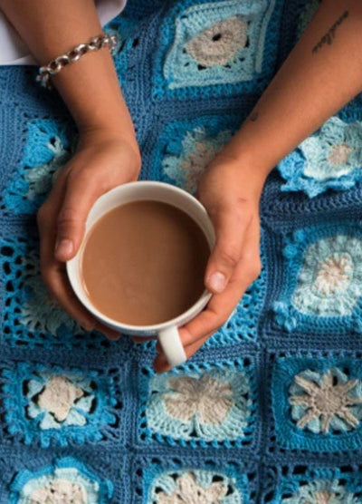 Hands holding a mug of coffee rest on a blue crocheted blanket. The blanket features intricate square patterns, creating a cozy, comforting scene.