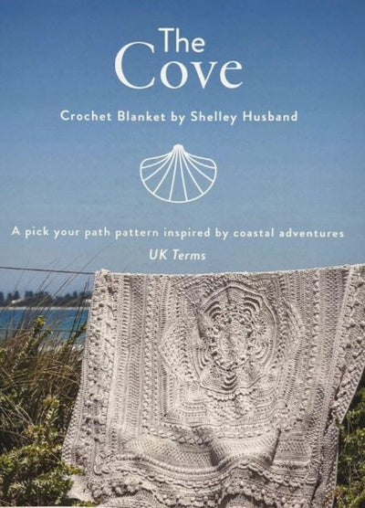 Cover image for a crochet blanket pattern book, "The Cove" by Shelley Husband. A detailed crochet blanket hangs outdoors, with a coastal backdrop.