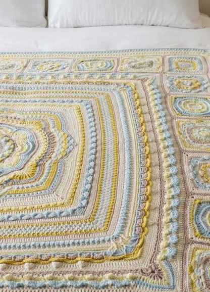 A crocheted blanket with intricate patterns in pastel colors like yellow, blue, and beige, neatly spread on a white bed, exuding warmth and coziness.