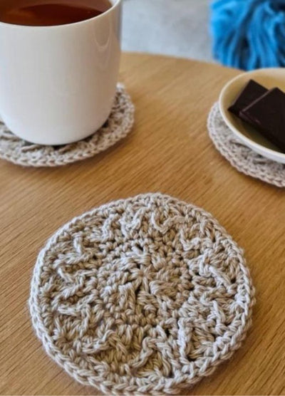 A white mug of tea and plate with chocolate sit on a wooden table, each on a beige crocheted coaster. Another coaster is in the foreground. Cozy and inviting.