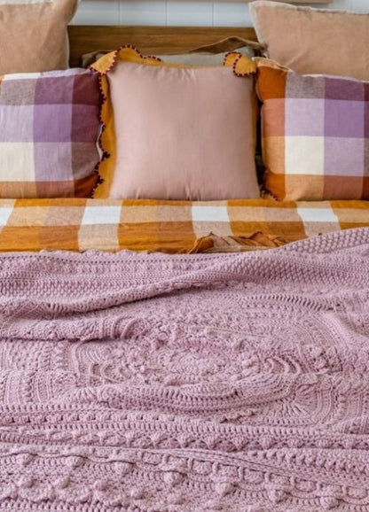 A cozy bed setup with soft, patterned pillows in muted tones, a checkered orange and purple bedspread, and a textured pink knitted blanket.