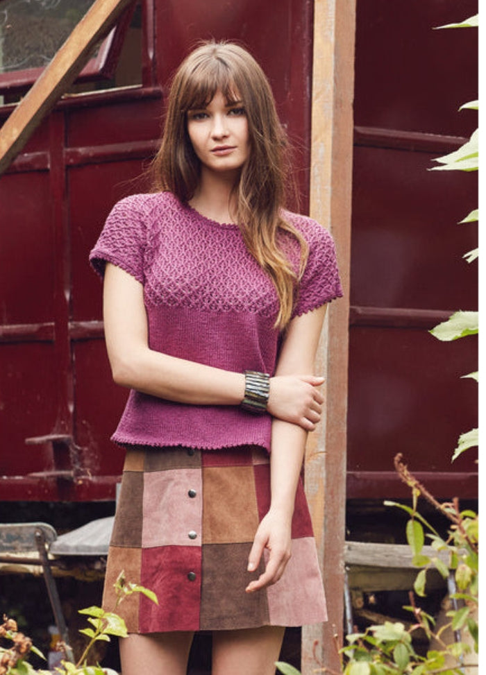 Model wearing the Sonam Tee from the Rowan Softyak DK Collection by Marie Wallin, a short-sleeved purple knit top paired with a colorful patchwork skirt, photographed against a bold red background with plants in the foreground.