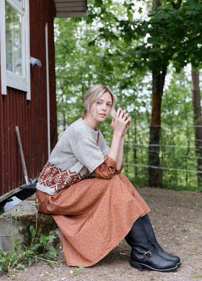 Saari Cardigan: light-colored knit with brown and white motifs at the waist, layered over an orange patterned dress and black boots outdoors.
