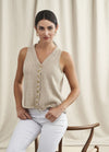 A woman in a beige knit sleeveless top and white pants stands against a light curtain background. She leans on a chair, exuding a calm and confident demeanor.