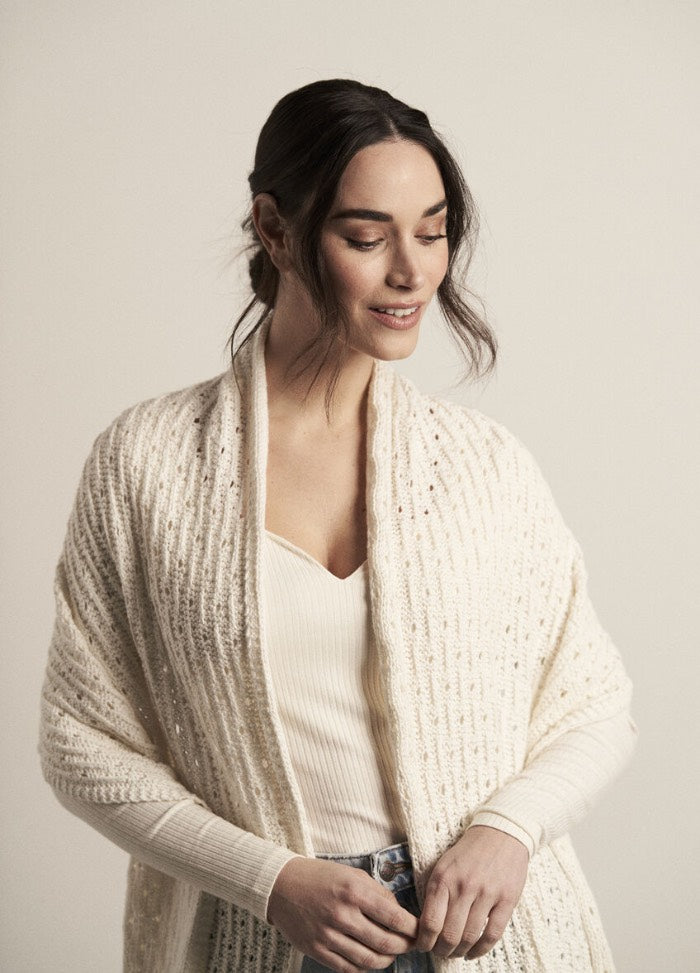Smiling woman wearing a cozy cream knit shawl and white top. She looks down softly, exuding warmth and serenity against a neutral background.