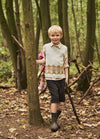 Young boy in outdoor setting stands beside a tree, holding a stick. He wears a light sweater vest and shorts, conveying an adventurous and playful mood.
