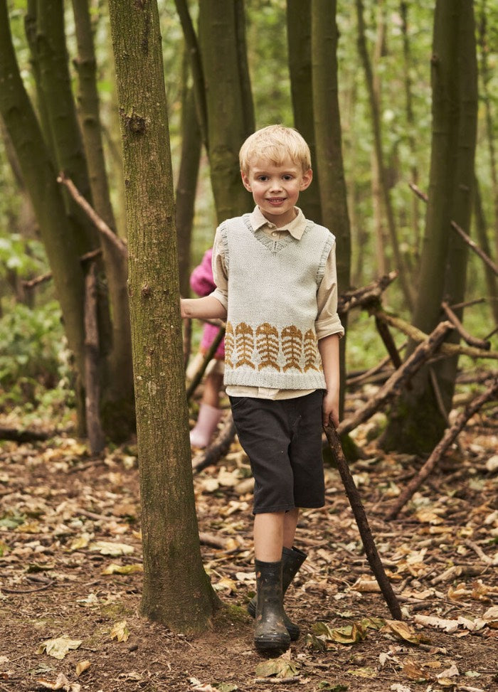 Young boy in outdoor setting stands beside a tree, holding a stick. He wears a light sweater vest and shorts, conveying an adventurous and playful mood.
