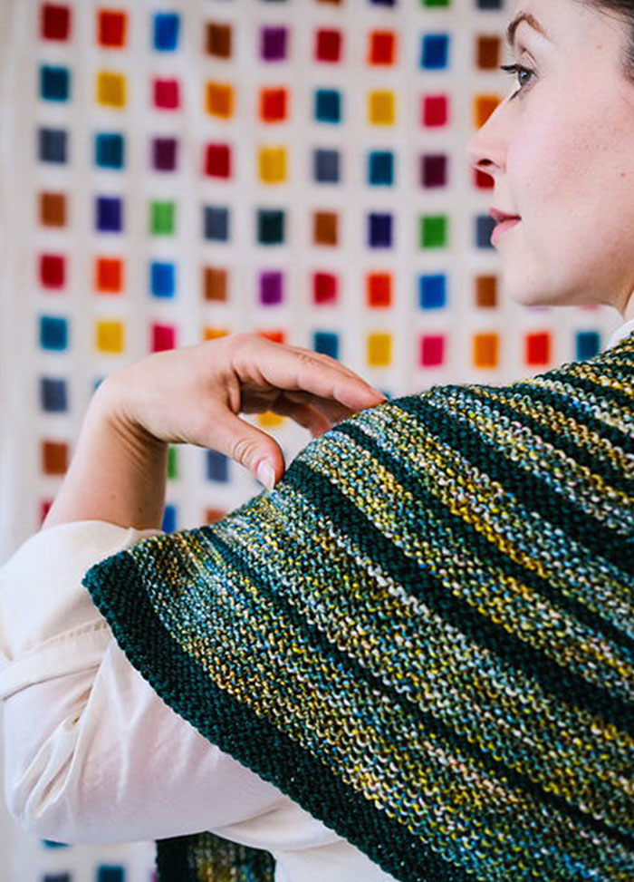 Back view of a striped triangular Saturn Shawl in green, yellow, and blue, worn against a colourful grid wall.