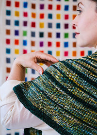 Back view of a striped triangular Saturn Shawl in green, yellow, and blue, worn against a colourful grid wall.
