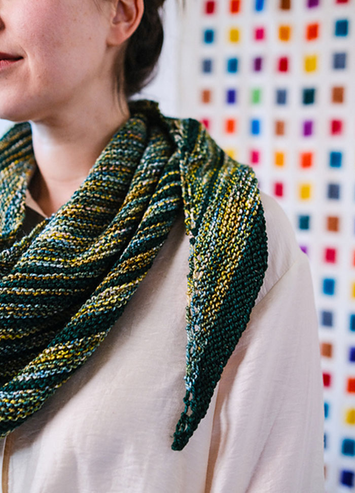 Striped triangular Saturn Shawl in green, yellow, and blue, draped around shoulders, shown against a colourful grid wall.