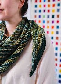Striped triangular Saturn Shawl in green, yellow, and blue, draped around shoulders, shown against a colourful grid wall.