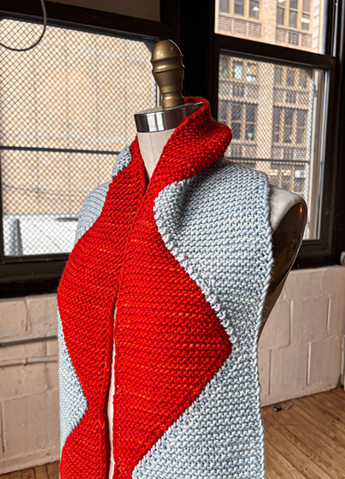 Sawtooth Scarf on a mannequin, featuring a bold zigzag pattern in alternating red and light blue sections, set against an urban indoor backdrop.