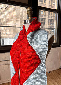 Sawtooth Scarf on a mannequin, featuring a bold zigzag pattern in alternating red and light blue sections, set against an urban indoor backdrop.