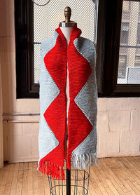 Sawtooth Scarf on a mannequin, featuring red and light blue zigzag stripes with light blue fringe, displayed indoors with large windows and a brick wall.