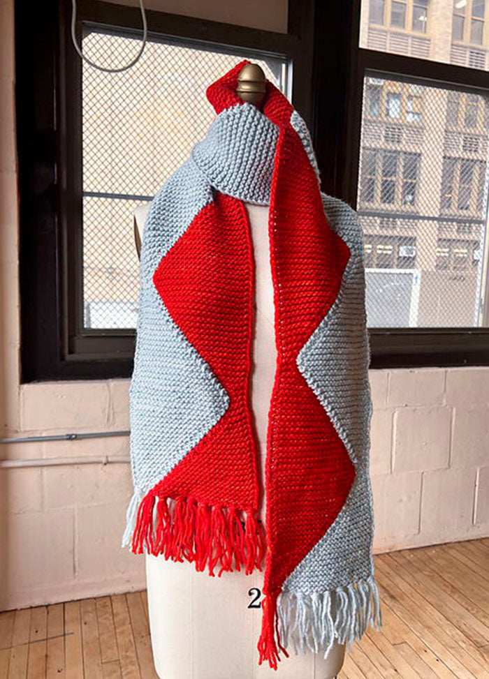Sawtooth Scarf on a mannequin, light gray with a bold red zigzag center and matching fringe, set against an urban indoor backdrop.