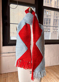 Sawtooth Scarf on a mannequin, light gray with a bold red zigzag center and matching fringe, set against an urban indoor backdrop.