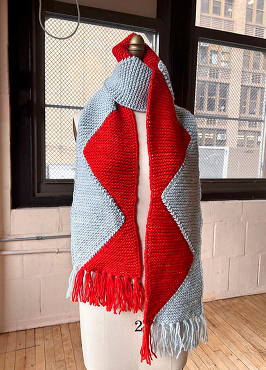 Sawtooth Scarf on a mannequin, light gray with a bold red zigzag center and matching fringe, set against an urban indoor backdrop.