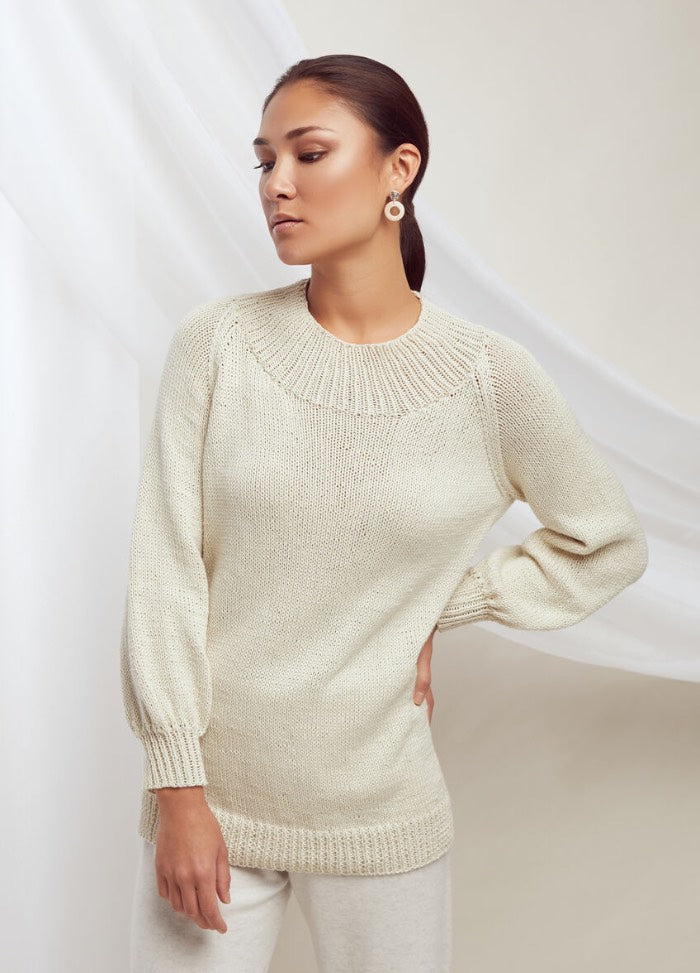 A person wearing a cream knit sweater and white pants stands against a soft beige background. They look thoughtful, exuding a calm, elegant vibe.