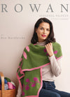 Woman in a turtleneck wears a green and pink knitted shawl with floral patterns. She is seated beside a basket of colorful yarn. The mood is cozy and elegant.