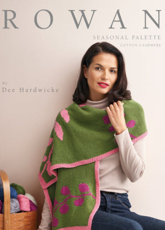 Woman in a turtleneck wears a green and pink knitted shawl with floral patterns. She is seated beside a basket of colorful yarn. The mood is cozy and elegant.