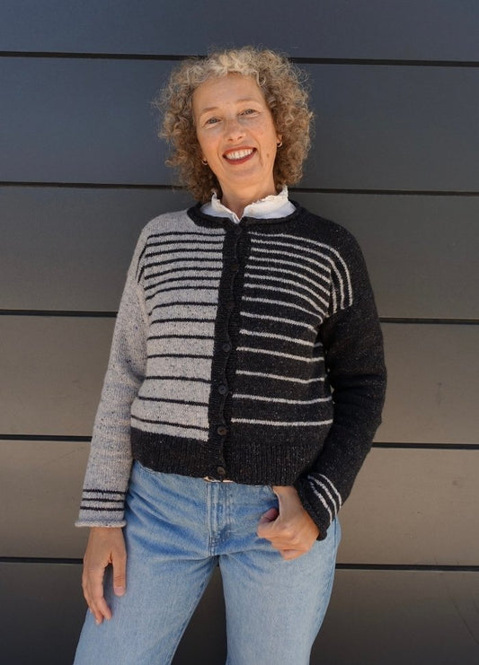 Smiling person with curly hair wears a striped, black and gray cardigan and light jeans against a dark, horizontally paneled background. Casual and cheerful mood.