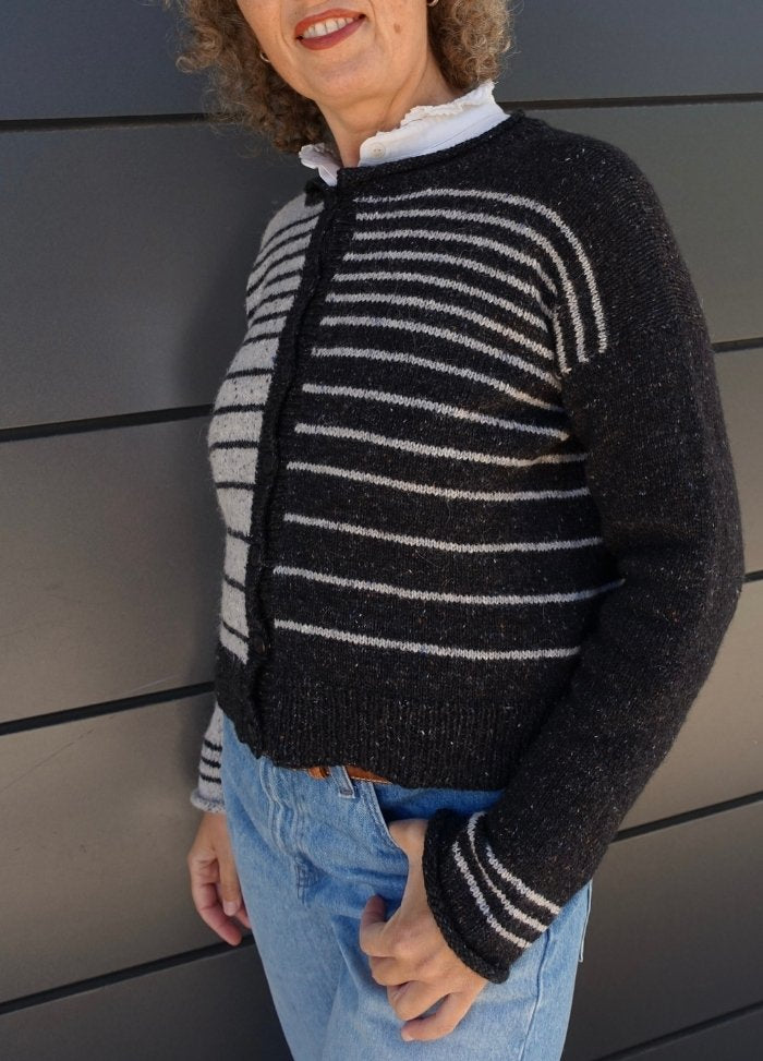 A person with curly hair wears a striped black and gray cardigan over a white shirt and blue jeans, standing against a dark paneled wall.