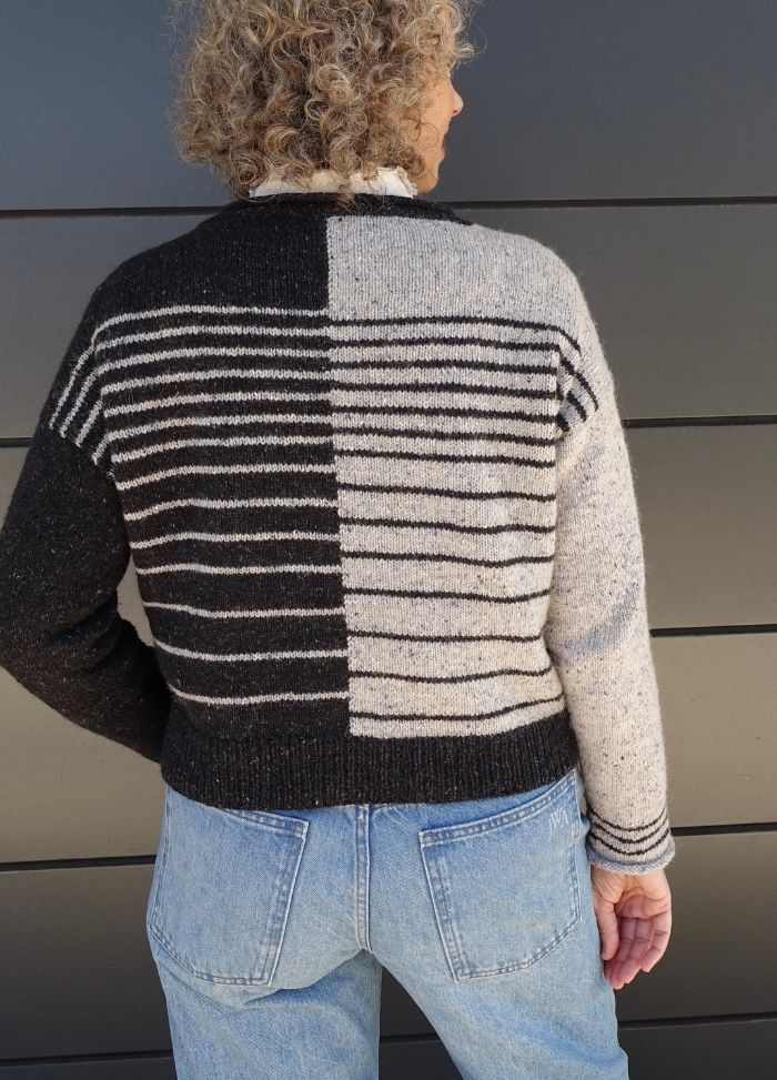 A person with curly hair wears a sweater with contrasting black and gray striped patterns, paired with blue jeans, standing against a dark background.