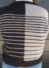 Back view of a knitted sweater with a split design: black with white stripes on the left, white with black stripes on the right, worn with jeans.