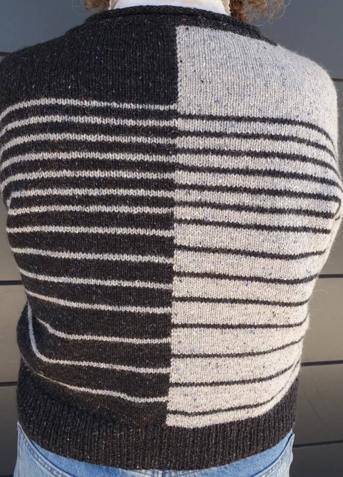 Back view of a knitted sweater with a split design: black with white stripes on the left, white with black stripes on the right, worn with jeans.