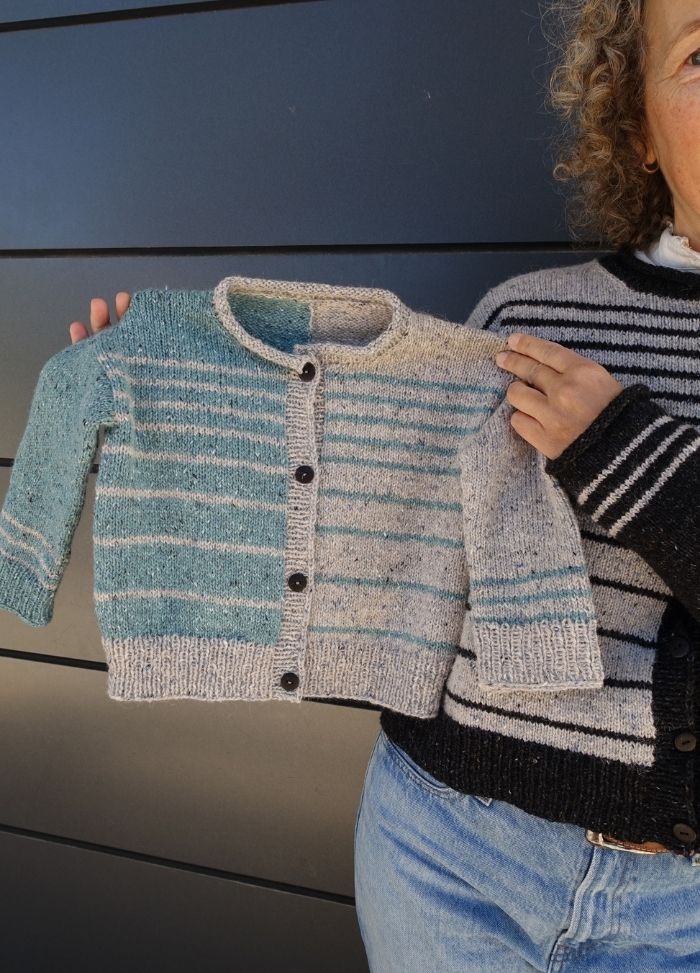 A person holds a small, hand-knit cardigan with turquoise and grey stripes, matching their own similar sweater. The scene conveys warmth and creativity.