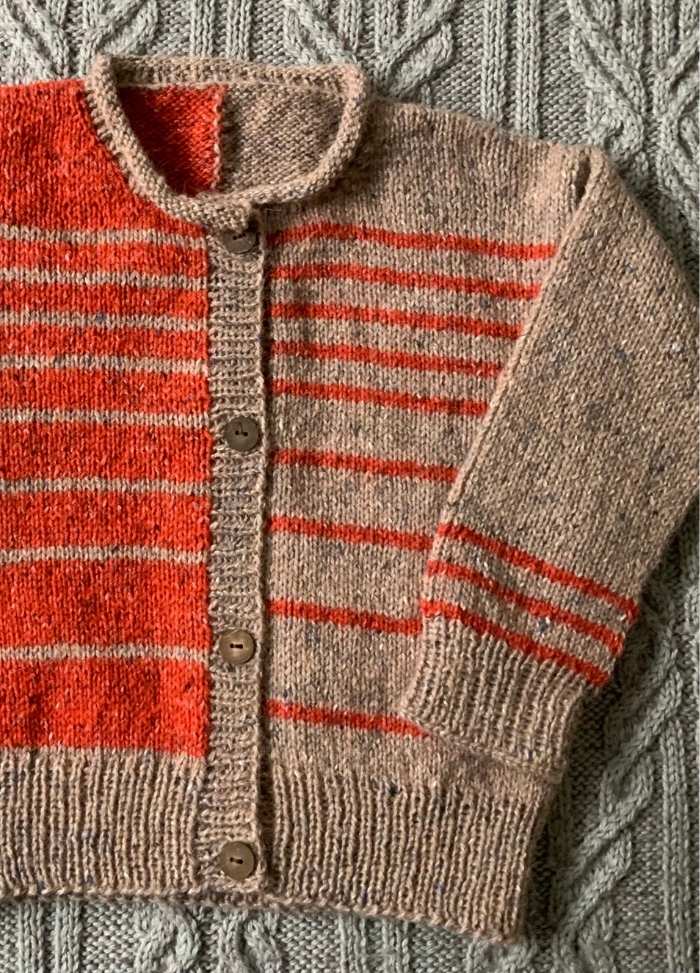 Hand-knitted cardigan in beige and orange stripes with brown buttons, laid on a textured gray cable-knit background. Cozy and warm vibe.