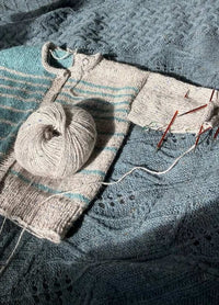 A partially knitted sweater with light gray and turquoise stripes and red knitting needles rests on a textured blue blanket, conveying a cozy, creative atmosphere.