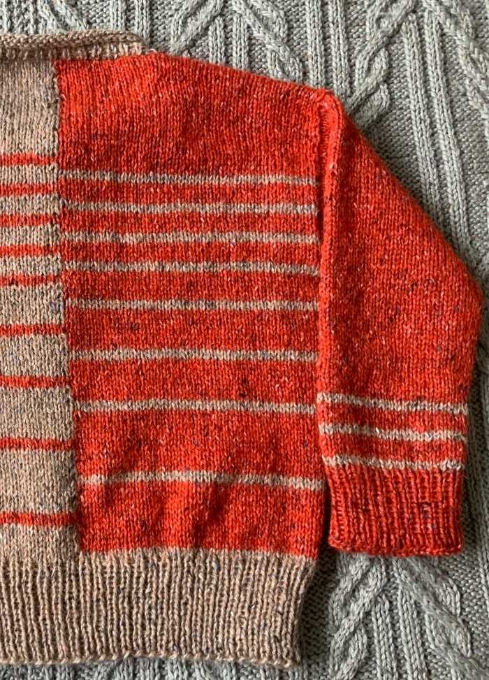 Close-up of a knitted sweater with orange and beige stripes on a gray, textured cable-knit background. The colors are vibrant and warm.