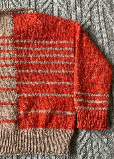 Close-up of a knitted sweater with orange and beige stripes on a gray, textured cable-knit background. The colors are vibrant and warm.