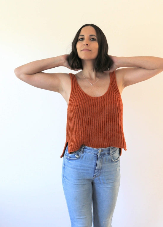 A person poses confidently in a sleeveless rust-orange knit top and light blue jeans against a plain white background, conveying a relaxed, casual vibe.