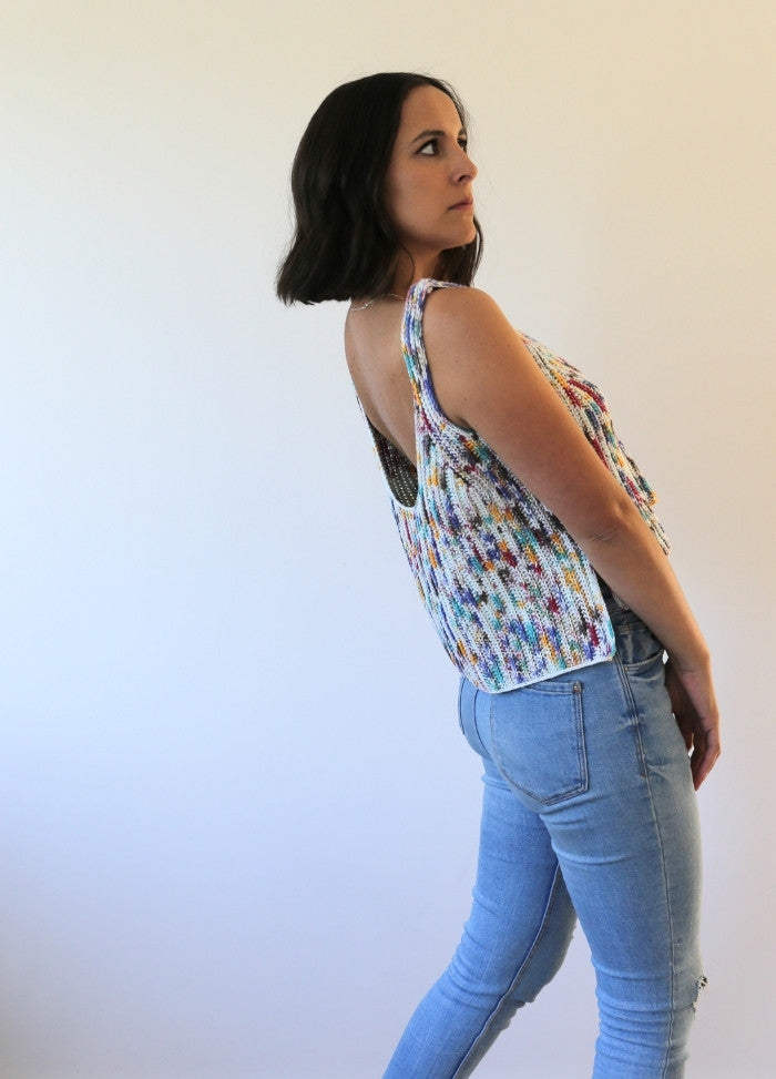 A woman with dark hair stands sideways, wearing a colorful knit top and blue jeans. She looks thoughtfully to the side against a plain background.