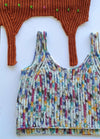 Knitted colorful crop top with blue, red, yellow, and black speckles on white. Above, a brown crocheted piece with green stitch markers. Playful and crafty.