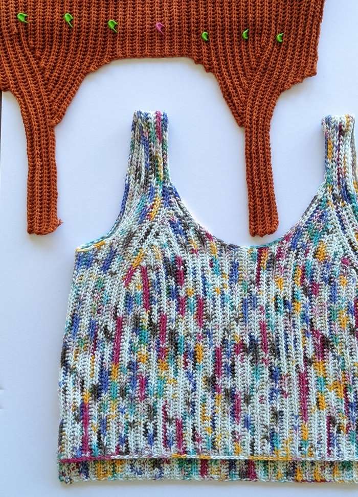 Knitted colorful crop top with blue, red, yellow, and black speckles on white. Above, a brown crocheted piece with green stitch markers. Playful and crafty.